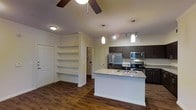 This single-floor space showcases an open-concept layout that seamlessly connects the living area to the kitchen. The kitchen is a standout feature, equipped with stainless steel appliances including a French door refrigerator, range, and built-in microwave. Dark espresso cabinetry provides strong visual contrast against the light granite countertops, which extend across a central island that also functions as a breakfast bar with an undermount sink. Pendant lights hang above the island, complementing the track lighting and ceiling fan that illuminate the space. The flooring throughout is rich, dark hardwood, adding warmth and continuity. On the living side, a built-in shelving unit with multiple open shelves offers practical storage and display space, flanked by a large closet door. A doorway leads further into the unit, hinting at additional rooms beyond the main living area. The overall ambiance is modern and functional, with a clean, neutral color palette accented by dark tones that give the space a contemporary, polished character.