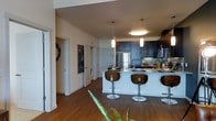 This single-floor residence showcases a thoughtfully designed open-concept layout that seamlessly connects the kitchen and living areas. The kitchen serves as the heart of the space, featuring rich dark cabinetry that extends to the upper walls, offering generous storage alongside a clean, contemporary aesthetic. A large central island with a light stone countertop provides ample workspace and doubles as a casual dining area, complemented by sleek modern bar stools with curved wooden backs. Stainless steel appliances, including a full-size refrigerator, add a polished, professional touch, while a tile backsplash introduces subtle texture to the design. Overhead, a trio of pendant lights — a mix of cylindrical and disc styles — cast warm, focused illumination over the island and surrounding space. Warm hardwood flooring flows throughout, grounding the space with a natural richness that contrasts beautifully with the lighter walls and cabinetry. Hallways branch off toward additional rooms, with glimpses of a washer and dryer unit visible, indicating in-unit laundry convenience. The high ceilings contribute to an airy, open feel, while the overall design balances modern sophistication with everyday functionality.