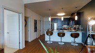 This single-floor residence showcases a thoughtfully designed open-concept layout that seamlessly connects the kitchen and living areas. The kitchen serves as the heart of the space, featuring rich dark cabinetry that extends to the upper walls, offering generous storage alongside a clean, contemporary aesthetic. A large central island with a light stone countertop provides ample workspace and doubles as a casual dining area, complemented by sleek modern bar stools with curved wooden backs. Stainless steel appliances, including a full-size refrigerator, add a polished, professional touch, while a tile backsplash introduces subtle texture to the design. Overhead, a trio of pendant lights — a mix of cylindrical and disc styles — cast warm, focused illumination over the island and surrounding space. Warm hardwood flooring flows throughout, grounding the space with a natural richness that contrasts beautifully with the lighter walls and cabinetry. Hallways branch off toward additional rooms, with glimpses of a washer and dryer unit visible, indicating in-unit laundry convenience. The high ceilings contribute to an airy, open feel, while the overall design balances modern sophistication with everyday functionality.