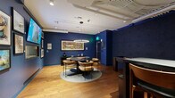 Located at 2512 W Colfax Ave in Denver, Colorado, this thoughtfully designed amenity lounge offers a vibrant and engaging social environment. The space is defined by its striking deep navy blue walls, which create a bold, immersive atmosphere complemented by warm hardwood flooring throughout.

The room is organized into distinct activity zones. At its center, a circular dining table surrounded by plush, patterned upholstered chairs serves as the primary gathering area, accented by a modern circular pendant light suspended overhead. A dedicated game area features a full shuffleboard table positioned against a dark textured accent wall, adding a playful and recreational dimension to the space.

A bar-height table with leather-seated stools provides an additional seating option, alongside a built-in counter area with open shelving displaying decorative objects. One wall is adorned with an extensive curated gallery of framed artwork and prints, alongside a large display screen, giving the lounge a distinct cultural and creative character. An enclosed aquarium is also integrated into the wall display, adding a unique and lively visual element.

Exposed ductwork on the white ceiling contributes an industrial aesthetic, while recessed and track lighting ensures the space is well-lit. The overall ambiance is sophisticated yet social, designed for both relaxed gatherings and recreational activities.