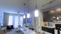 This single-floor residence offers a thoughtfully designed open-concept layout that seamlessly connects the kitchen and living areas into one cohesive and inviting space. The kitchen is a standout feature, showcasing granite countertops, a stainless steel sink, and dark cabinetry that provides a sophisticated contrast to the light-toned surfaces. A trio of elegant pendant lights hang above the kitchen island, casting warm illumination over the prep and dining space, while a track lighting fixture adds further versatility. The living area is comfortably appointed with a plush sofa, a low-profile coffee table, and a decorative area rug. A ceiling fan with a brushed metal finish keeps the space comfortable year-round. Large windows fitted with white blinds invite abundant natural light throughout the day, contributing to the bright and open ambiance. Neutral wall tones serve as a clean backdrop, accented by contemporary artwork that adds a touch of color and personality. A centrally located interior door provides access to additional areas of the residence, completing this well-balanced and modern living environment.