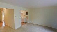This two-floor residential space offers a straightforward and functional layout finished in soft, neutral tones that create a calm and welcoming atmosphere throughout. The main living area is carpeted in a light beige, providing a comfortable and quiet underfoot experience, while the walls are painted in a clean off-white that keeps the space feeling bright and open. An open passageway connects the living area directly to the kitchen, allowing for an easy flow between the two spaces. The kitchen features white cabinetry — both upper and lower — along with a sink, countertop workspace, and wood-style flooring that contrasts neatly with the carpeted areas. The cabinetry includes a decorative glass-panel upper cabinet, adding a subtle design touch to an otherwise practical space. The overall aesthetic is clean and understated, with a simple, move-in-ready feel. The two-floor layout offers additional living space beyond what is visible, providing room for bedrooms, bathrooms, or supplementary areas on the second level. The neutral finishes and practical design make this a versatile space well-suited to a variety of living arrangements.