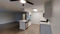This single-floor space is designed with an open-concept layout that maximizes functionality and flow. The kitchen serves as the centerpiece, featuring a central island with a built-in sink, sleek white cabinetry, and crisp white countertops that lend a clean, modern aesthetic. Stainless steel appliances — including a dishwasher, gas range, and microwave — provide a well-equipped cooking environment. A skylight panel above the kitchen floods the workspace with natural light, complementing the recessed and flush-mount lighting throughout. The kitchen transitions seamlessly into a generously sized living area, unified by warm wood-look flooring that runs throughout the space. The living area features a ceiling fan, adding both style and practicality. Soft, cool-toned accent walls in muted blue-gray contrast tastefully with the bright white trim and cabinetry, creating a calm and contemporary ambiance. The open sightlines between the kitchen and living area make the space feel expansive and inviting, ideal for both everyday living and entertaining. The overall design balances modern finishes with a welcoming, airy atmosphere.
