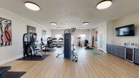 This fitness center is a single-floor facility designed to support a full spectrum of workout routines. The space features light wood-style flooring throughout, white walls, and flush-mount circular ceiling lights that provide consistent, even illumination across the entire area. Large windows allow natural light to complement the artificial lighting, contributing to an open and energizing atmosphere.

The strength training section is well-appointed with multiple cable and resistance machines, a multi-function weight station, a dedicated dumbbell rack, and a selection of medicine balls on a wall-mounted rack. A central cable tower serves as a focal point of the space, offering versatile exercise options. Rubber floor mats are placed under each piece of equipment for stability and floor protection.

The cardio zone includes treadmills, an elliptical trainer, a stationary recumbent bike, and a stair-stepper, offering varied options for aerobic training. A designated stretching and functional training area features yoga mats, aerobic step platforms, and stability balls, providing space for warm-ups and bodyweight exercises.

A wall-mounted television, a built-in cabinetry station, and motivational typography artwork displayed throughout add to the facility's functional and encouraging environment. A glass-paneled door offers direct access to the outdoors, enhancing ventilation and connectivity to the surrounding area.