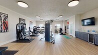 This fitness center is a single-floor facility designed to support a full spectrum of workout routines. The space features light wood-style flooring throughout, white walls, and flush-mount circular ceiling lights that provide consistent, even illumination across the entire area. Large windows allow natural light to complement the artificial lighting, contributing to an open and energizing atmosphere.

The strength training section is well-appointed with multiple cable and resistance machines, a multi-function weight station, a dedicated dumbbell rack, and a selection of medicine balls on a wall-mounted rack. A central cable tower serves as a focal point of the space, offering versatile exercise options. Rubber floor mats are placed under each piece of equipment for stability and floor protection.

The cardio zone includes treadmills, an elliptical trainer, a stationary recumbent bike, and a stair-stepper, offering varied options for aerobic training. A designated stretching and functional training area features yoga mats, aerobic step platforms, and stability balls, providing space for warm-ups and bodyweight exercises.

A wall-mounted television, a built-in cabinetry station, and motivational typography artwork displayed throughout add to the facility's functional and encouraging environment. A glass-paneled door offers direct access to the outdoors, enhancing ventilation and connectivity to the surrounding area.
