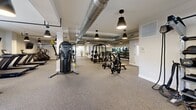 This state-of-the-art fitness facility, located at 150 Broad St in Windsor, CT, offers a spacious 1,840 square foot workout area on a single floor. The center boasts an industrial-chic design with exposed ductwork, pendant lighting, and a neutral color palette. Multiple exercise zones cater to various fitness needs, including a cardio area with treadmills and cycling equipment, a strength training section with weight machines and free weights, and a functional fitness space with suspension trainers and plyometric equipment. Large windows provide natural light, while potted plants add a touch of greenery. A feature wall with a chalkboard allows for workout tracking or motivational messages. The facility is equipped with modern amenities such as wall-mounted TVs and water fountains. The open floor plan and strategic equipment placement ensure ample space for movement and comfortable workouts. This comprehensive fitness center is designed to accommodate diverse exercise routines and fitness levels in a clean, well-maintained environment.