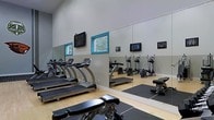 This fitness center is designed to support a well-rounded workout experience within a bright, open layout. The space features soaring ceilings that contribute to an airy, open atmosphere, while light-toned hardwood-style flooring runs throughout, lending a clean and polished look. A row of treadmills lines one wall, providing ample cardio options for users, while elliptical trainers and additional cardiovascular equipment are positioned nearby to offer variety. A full-length wall mirror spans a significant portion of the room, enhancing the sense of space and allowing users to monitor their form during exercise. Weight benches and free weights are also available, supporting strength training alongside the cardio offerings. Mounted flat-screen televisions are positioned at eye level, providing entertainment during workouts. Natural light filters into the space through teal-framed windows, adding a distinctive pop of color to the otherwise neutral gray and white walls. Rubber floor mats are placed in the free weights area to protect the flooring and provide stability. Overall, the fitness center is thoughtfully arranged to maximize functionality while maintaining a clean, modern aesthetic.