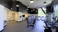 This contemporary fitness center offers a well-appointed workout environment designed to meet a wide range of exercise needs. The space features soaring ceilings finished in a bold black tone, creating a striking contrast against the white lower walls and light-colored flooring. A generous array of pendant lights hang at varying heights throughout the room, providing bright, even illumination across the entire floor.

The workout area is thoughtfully organized, with a dedicated strength training zone housing multiple cable and resistance machines suited for a full-body workout. A row of treadmills lines the perimeter near a large floor-to-ceiling window, which floods the space with natural light and offers a refreshing view of lush greenery outside. A wall-mounted flat-screen television is positioned for easy viewing during cardio sessions.

Additional equipment, including free weights and benches, is neatly arranged toward the back of the room. Built-in shelving along the wall provides convenient storage for accessories and essentials. The rubber flooring throughout the space ensures both safety and durability. The overall ambiance is clean, modern, and energizing, making this fitness center a functional and visually appealing facility for daily use.