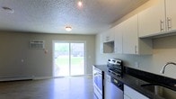 This single-floor space showcases a combined kitchen and living area designed for functional, everyday living. The kitchen is equipped with stainless steel appliances, including an electric range, and features white cabinetry with sleek bar-pull hardware providing ample storage. Dark countertops offer a contrasting accent to the bright, white cabinetry above. Light wood-style laminate flooring runs throughout, adding warmth to the neutral grey walls.

A textured popcorn ceiling adds depth overhead, while flush-mount ceiling lighting ensures the interior remains well-lit. A standout feature of the space is the full-width sliding glass patio door, which floods the area with natural light and provides direct access to an outdoor patio and yard area. The view through the door reveals a fenced backyard, offering a sense of privacy.

A wall-mounted air conditioning unit is positioned near the patio door, ensuring climate control year-round. The open layout between the kitchen and the adjoining area creates a practical and connected flow, making this a comfortable and efficiently designed single-level space.