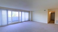 This single-floor space offers a clean, functional layout designed to maximize comfort and natural light. The main living area is spacious and open, finished with soft, neutral-toned wall-to-wall carpeting that adds warmth underfoot. Large sliding glass doors dominate one wall, fitted with vertical blinds that allow for flexible light control. These doors open to a private balcony, inviting natural light deep into the interior while offering pleasant outdoor views of surrounding greenery. The walls are finished in a soft, warm off-white tone that keeps the space feeling bright and airy. Ceiling-mounted HVAC vents are thoughtfully integrated throughout, ensuring efficient climate control across the living area. The room's generous proportions provide ample space for a variety of furniture arrangements, making it highly versatile. The overall ambiance is calm and understated, with a neutral palette that serves as an excellent canvas for personalization. The combination of abundant natural light, outdoor access, and open layout makes this a welcoming and practical living environment.