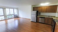 This single-floor space offers an open, airy layout that seamlessly connects the living area and kitchen, creating a functional and inviting environment. Rich hardwood flooring extends throughout, adding warmth and continuity to the space.

The kitchen is outfitted with warm-toned wood cabinetry, granite countertops, and stainless steel appliances including a full-size refrigerator and dishwasher, providing both style and practicality. The kitchen flows directly into the expansive living area, making the space well-suited for everyday living and entertaining.

The living area benefits from generous natural light streaming in through a large sliding glass door, which also provides access to an outdoor area and frames views of the surrounding environment. A wall-mounted air conditioning unit ensures year-round comfort. The clean white walls and open floor plan contribute to a bright, versatile atmosphere that can be easily personalized. Overall, the space strikes a balance between functional design and comfortable living.