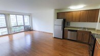 This single-floor space offers an open, airy layout that seamlessly connects the living area and kitchen, creating a functional and inviting environment. Rich hardwood flooring extends throughout, adding warmth and continuity to the space.

The kitchen is outfitted with warm-toned wood cabinetry, granite countertops, and stainless steel appliances including a full-size refrigerator and dishwasher, providing both style and practicality. The kitchen flows directly into the expansive living area, making the space well-suited for everyday living and entertaining.

The living area benefits from generous natural light streaming in through a large sliding glass door, which also provides access to an outdoor area and frames views of the surrounding environment. A wall-mounted air conditioning unit ensures year-round comfort. The clean white walls and open floor plan contribute to a bright, versatile atmosphere that can be easily personalized. Overall, the space strikes a balance between functional design and comfortable living.
