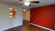 Located at 342 Riverwood Dr. in Crestview, Florida, this single-story home offers a welcoming and practical layout designed for comfortable everyday living. The main living area immediately draws attention with its rich, dark hardwood-style flooring that extends throughout the open space, providing a warm and cohesive foundation. A striking deep-red accent wall adds a bold design element, creating a focal point that anchors the room with color and character. Ceiling fans with integrated lighting fixtures are present in the living spaces, contributing to both comfort and illumination. The open-concept design connects the living area seamlessly to the kitchen, which is accessible via a convenient pass-through counter. The kitchen features wooden cabinetry and appears well-appointed for everyday use. White baseboards frame the flooring throughout, offering a clean contrast to the walls and flooring. The overall ambiance is inviting and functional, blending practical design choices with a few standout decorative touches. Neutral wall tones in the main living space complement the bolder accent wall, creating a balanced and versatile interior. This single-floor layout ensures easy accessibility and a straightforward flow between spaces.