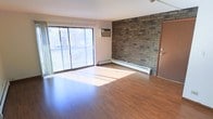 This single-floor space offers a clean, functional layout with a welcoming ambiance defined by a blend of classic and practical design elements. The main living area is anchored by warm hardwood-style flooring that extends throughout, lending a cohesive and inviting feel underfoot. A striking full-height exposed brick accent wall serves as the room's focal point, adding texture, character, and a timeless architectural detail that contrasts beautifully with the smooth, neutral-toned walls. Natural light pours generously into the space through a large sliding glass door, which opens directly onto an outdoor balcony framed by a white railing. The balcony overlooks a lush, tree-lined exterior, providing a pleasant connection to the outdoors. A wall-mounted air conditioning unit ensures year-round comfort, while baseboard heating runs along the perimeter of the room, offering an efficient and unobtrusive heating solution. The overall layout feels open and airy, with ample room to furnish and personalize. The space combines durable, low-maintenance finishes with architectural charm, making it a practical yet characterful place to call home.
