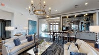 This thoughtfully designed lobby offers a seamless blend of comfort and sophistication across a single, open floor. Upon entry, the space immediately impresses with a striking wrought-iron candle-style chandelier that anchors the seating area below. Plush neutral sofas and accent chairs are arranged around a glass-top coffee table, creating an inviting lounge environment accented by bold blue throw pillows and decorative sculptures.

A large, vibrant blue abstract painting serves as a prominent focal point on one wall, adding an artistic energy to the space. The flooring features rich dark hardwood, complemented by a patterned area rug that defines the lounge zone.

Deeper into the lobby, a dining area with high-top tables and dark wood bar-height chairs offers a more casual gathering space. Adjacent to this, a well-appointed bar area is framed by an exposed stone accent wall, backlit shelving displaying glassware and bottles, and a mirrored backdrop — lending a refined, club-like ambiance.

Recessed lighting and coffered ceiling details in the bar zone add architectural interest, while large windows and glass doors at the rear draw in natural light and provide views to an outdoor area, enhancing the overall sense of openness and connectivity throughout the lobby.