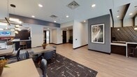 This sophisticated single-floor amenity lounge is designed to offer a variety of social and leisure experiences within a cohesive, modern aesthetic. Light wood-plank flooring flows throughout, unifying the distinct zones and complementing the warm, contemporary design palette.

The centerpiece of the main lounge is a dramatic floor-to-ceiling stone tile fireplace wall with a linear gas fireplace, flanked by large sectional sofas, upholstered ottomans, and accent chairs arranged atop a patterned area rug. Oversized windows and sliding glass doors flood the space with natural light and provide direct access to an outdoor terrace furnished with lounge seating.

A dedicated bar area features dark cabinetry, tile-wrapped islands with white marble countertops, pendant lighting, and built-in display shelving — ideal for entertaining. Adjacent to this is a fully equipped catering kitchen with stainless steel appliances, a large refrigerator, a decorative geometric-patterned accent wall, and a bar-height island with wood stools.

A dining and lounge nook with booth-style seating, pedestal tables, and a wall-mounted TV opens to additional terrace access via sliding glass doors. The entry area features curated wall art, a patterned area rug, and a dining table — serving as a welcoming transition into the broader space. Recessed lighting throughout ensures a warm, well-lit ambiance across all zones.