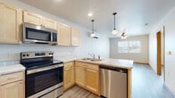 This single-floor unit offers a thoughtfully designed layout that balances functionality and comfort throughout. The open-concept kitchen and living area form the heart of the space, anchored by light maple cabinetry, stainless steel appliances including a refrigerator, electric range, built-in microwave, and dishwasher, as well as light-toned countertops. A peninsula with pendant lighting overhead serves as a natural divider between the kitchen and the adjoining living area, which is illuminated by a ceiling fan with integrated lighting and benefits from natural light through a large window. Light gray wood-look flooring flows consistently throughout the entire unit, lending a cohesive and modern feel. The bedrooms are generously proportioned, each featuring ceiling fans, natural light from windows with exterior views, and warm wood baseboards that complement the flooring. One bedroom is additionally equipped with a wall-mounted mini-split HVAC unit for individualized climate control. The bathroom is notably designed with accessibility in mind, featuring a walk-in shower with stainless steel grab bars and a built-in seat, offering both safety and practicality. The overall ambiance of the space is clean, bright, and move-in ready, with a neutral color palette that provides a versatile backdrop throughout.