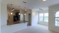 This two-floor residence offers a thoughtfully designed open-concept layout that seamlessly connects the kitchen and living areas. The kitchen is equipped with dark cabinetry, dark granite-style countertops, a built-in sink, and a black refrigerator. A breakfast bar extends the counter space, ideal for casual dining or entertaining. Pendant lighting hangs above the bar, adding a warm, focused glow to the kitchen zone. Adjacent to the kitchen, a built-in shelving unit provides convenient storage and display space, adding both function and character to the main living area. The living room is finished with plush wall-to-wall carpeting that lends a comfortable, inviting feel to the space. A large multi-pane window floods the room with natural light and offers views of the surrounding greenery outside. Track lighting overhead in the living area allows for flexible and customizable illumination. The clean white walls and neutral tones throughout create a bright, airy ambiance. Ceiling vents are thoughtfully positioned to ensure efficient climate control across the space. With its well-considered design and functional layout spread across two floors, this residence offers a welcoming and versatile living environment.