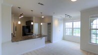This two-floor residence offers a thoughtfully designed open-concept layout that seamlessly connects the kitchen and living areas. The kitchen is equipped with dark cabinetry, dark granite-style countertops, a built-in sink, and a black refrigerator. A breakfast bar extends the counter space, ideal for casual dining or entertaining. Pendant lighting hangs above the bar, adding a warm, focused glow to the kitchen zone. Adjacent to the kitchen, a built-in shelving unit provides convenient storage and display space, adding both function and character to the main living area. The living room is finished with plush wall-to-wall carpeting that lends a comfortable, inviting feel to the space. A large multi-pane window floods the room with natural light and offers views of the surrounding greenery outside. Track lighting overhead in the living area allows for flexible and customizable illumination. The clean white walls and neutral tones throughout create a bright, airy ambiance. Ceiling vents are thoughtfully positioned to ensure efficient climate control across the space. With its well-considered design and functional layout spread across two floors, this residence offers a welcoming and versatile living environment.