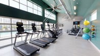 This single-floor fitness center is designed to deliver a comprehensive workout experience in a bright, open environment. The space is defined by soaring vaulted ceilings painted in a bold teal and green palette, which lend an energetic and motivating atmosphere throughout. Expansive floor-to-ceiling windows line one wall, flooding the interior with abundant natural light while offering views of an outdoor swimming pool — a refreshing backdrop for any workout session. Large ceiling fans are suspended overhead, complementing the exposed ductwork that reinforces the facility's modern industrial aesthetic. The light wood-style flooring enhances the clean, contemporary look of the space. The cardio zone is generously appointed with a row of Matrix treadmills, elliptical cross-trainers, and stationary cycling bikes, each equipped with individual screens. A dedicated free weights and strength training area features a dumbbell rack, a wall-mounted TV, and various benches. The functional training section offers a power tower, medicine balls, resistance bands, yoga mats, and a suspension training system mounted to the wall, providing options for a full-body workout. A water fountain station and additional amenities are accessible near the entry, completing this well-rounded and thoughtfully outfitted fitness facility.