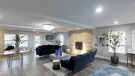 Located at 320 Waterford Ct in Lafayette, Indiana, this single-floor space offers a thoughtfully designed interior that blends modern aesthetics with comfortable, functional living. The centerpiece of the main lounge area is a striking floor-to-ceiling stacked stone fireplace, which anchors the room and adds a warm, textural contrast to the otherwise sleek surroundings. The space is furnished with deep navy blue sofas and contemporary accent pieces, including a metallic coffee table and coordinating side stools, all arranged atop a layered area rug over smooth hardwood-style flooring. Recessed lighting and a classic flush-mount ceiling fixture provide even, inviting illumination throughout the room, while natural light filters in through windows dressed with clean white blinds. Soft gray walls and crisp white trim create a neutral, versatile backdrop that enhances the overall sense of openness. Curated wall art and lush indoor greenery add finishing touches that elevate the ambiance. A wall-mounted television and direct access to an exterior door further enhance the practicality and accessibility of this well-appointed space, making it equally suited for relaxed gatherings or everyday use.
