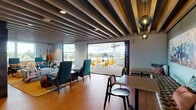 Located at 2869 NE Sandy Blvd in Portland, Oregon, this thoughtfully designed single-floor space blends mid-century modern aesthetics with contemporary functionality. A signature feature throughout is the striking exposed beam ceiling, finished in deep charcoal tones, which adds architectural character and visual depth to the open interior.

The main lounge area is anchored by warm wood-look tile flooring, an array of mid-century style seating, and a floor-to-ceiling glass fireplace set against a dramatic dark stone tile wall. A wall-mounted television makes the space ideal for gathering. Large windows and expansive folding glass doors flood the interior with natural light and connect seamlessly to an outdoor rooftop terrace, where views of the Portland cityscape and surrounding greenery can be enjoyed from colorful accent seating.

A dedicated kitchen area features sleek white cabinetry, modern appliances, and a clean, minimalist aesthetic with tile flooring and generous counter space. Nearby, a bar-height counter with stools and pendant lighting provides additional seating, while curated artwork — including vivid bird-themed prints — lends a creative, local character to the walls. The overall ambiance is vibrant yet welcoming, combining purposeful design with eclectic, colorful decor throughout.
