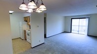 This single-floor space showcases an open-concept design that seamlessly connects the kitchen and living area. The kitchen is equipped with white appliances, including a freestanding gas range and a full-sized refrigerator, complemented by warm wood-toned cabinetry. Tile flooring in the kitchen transitions into plush wall-to-wall carpeting that extends throughout the spacious living area, adding comfort underfoot. A multi-bulb chandelier with classic bell-shaped shades serves as a central lighting fixture, providing ample illumination to the shared space. The living area is generously proportioned, featuring neutral white walls and dark baseboard trim that frame the room cleanly. A sliding glass door at the far end of the living space floods the interior with natural light and provides direct access to an outdoor balcony, offering views to the exterior. A hallway visible from the main living area leads to additional spaces, including what appears to be a closet with built-in storage. The overall layout is practical and functional, with a straightforward flow between rooms suited for everyday living.