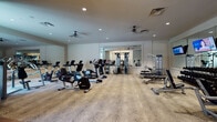 This fitness center is thoughtfully designed to offer a comprehensive workout environment within an elegantly appointed space. The main exercise area is generously sized, accommodating a full suite of Life Fitness cardio equipment, including treadmills, ellipticals, stationary bikes, and a recumbent bike. A multi-station cable machine anchors the strength training section, complemented by a full dumbbell rack, adjustable weight benches, and additional selectorized machines for targeted muscle training. Stability balls and accessories are neatly organized on a dedicated rack nearby. Large wall-mounted mirrors run the length of key sections, visually expanding the space and supporting proper form during workouts. Mounted flat-screen televisions provide entertainment throughout the session. Ceiling fans and recessed lighting ensure a comfortable, well-lit atmosphere. The distinctive patterned carpet — featuring a neutral, animal-print motif — adds a refined aesthetic touch throughout. A welcoming lounge area at the entry transitions from hardwood flooring to the main fitness space, furnished with upholstered accent chairs, a classic wooden side table, and a striking brass chandelier beneath soaring windows that invite natural light and garden views, creating a warm and inviting arrival experience.
