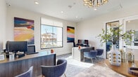 Located at 9944 Highway 6 in Missouri City, TX, this single-floor office space offers a thoughtfully designed and well-appointed environment for professional use. The layout seamlessly integrates a welcoming reception area with a functional workspace, creating a cohesive flow throughout.

Upon entry, a bright reception area greets visitors with warm stone-look tile flooring laid in a herringbone pattern, crisp white walls, and a stylish pedestal accent table. Comfortable gray lounge seating and framed colorful artwork contribute to an inviting, modern atmosphere. Large glass-paneled entry doors flood the space with natural light and provide views of the surrounding community.

The main office area features two sleek wood-tone workstations, each paired with contemporary blue upholstered guest chairs. Bold, large-scale abstract paintings in vibrant hues add a creative energy to the room, while a striking gold multi-ring chandelier serves as a distinguished focal point overhead. A generously sized window frames an outdoor view, further enhancing the bright and airy feel of the workspace.

Additional functional areas include a secondary workstation with a wall-mounted display screen, a frosted-glass interior door providing access to a private room, and French doors connecting to an adjacent space. Recessed lighting throughout ensures consistent, professional illumination across the entire office.