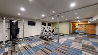 This fitness center is a comprehensively outfitted exercise space designed to support a variety of workout routines. The room is laid out to maximize usability, with distinct zones dedicated to different types of training. A generous selection of cardio equipment, including treadmills, elliptical trainers, and a recumbent bike, occupies a central area of the floor plan, each positioned for comfortable use and easy circulation around them.

Along one wall, a rack of dumbbells provides free weight options, while a dedicated corner area features colorful stability and medicine balls alongside additional functional fitness accessories, all set against a warm orange accent wall that adds a vibrant contrast to the otherwise neutral color palette. Wall-mounted flat-screen televisions are positioned throughout the space, allowing users to stay entertained during workouts.

The flooring consists of eye-catching modular carpet tiles in layered shades of blue, grey, and tan, adding visual interest underfoot. Recessed lighting illuminates the space evenly from above, and a wall-mounted climate control unit ensures a comfortable environment. A large mirror visually expands the room, while a bold entrance mat and blue entry door mark the main access point.