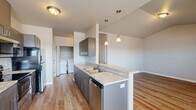 This thoughtfully laid-out 2-bedroom, 2-bathroom single-floor residence offers a functional and modern living experience. The open-concept kitchen and living area serves as the heart of the home, anchored by a central island with a double stainless steel sink and light granite-style countertops. Gray shaker-style cabinetry lines the kitchen, complemented by stainless steel appliances including a dishwasher. Pendant lighting above the island adds a contemporary touch, while natural light pours in through windows overlooking the surrounding views. In-unit washer and dryer connections are conveniently located nearby. Wood-look flooring flows consistently throughout the entire space, creating warmth and visual continuity. The primary bedroom features vaulted ceilings that lend an airy, spacious feel, along with a window for natural light. The second bedroom is generously proportioned and includes a closet with built-in shelving and hanging rods. Two full bathrooms serve the unit: one with a soaking tub and shower combination, and the other with a dedicated walk-in shower stall. Both bathrooms feature matching gray cabinetry with granite-style countertops and wood-look flooring. A hallway connects all rooms, completing this efficiently designed and move-in-ready residence.