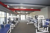 This fitness center offers a comprehensive workout environment designed to accommodate a wide range of exercise routines. The space is organized into distinct zones, with cardio equipment — including treadmills, stationary bikes, and elliptical machines — positioned near large, grid-paned windows that allow abundant natural light and offer views of the surrounding outdoor area. A row of strength training machines lines the interior, featuring a variety of cable and weight resistance stations suited for full-body workouts.

A defining architectural element is the exposed red ductwork running along the ceiling, set against a dark gray ceiling finish, which gives the space an industrial character. Flush-mounted fluorescent light panels provide consistent overhead illumination throughout. The flooring consists of durable, textured carpet tiles well-suited for a high-traffic fitness environment. Ceiling fans add to the ventilation and comfort of the space.

A wall-mounted television is positioned centrally, visible from multiple workout areas. A water fountain is accessible near the interior wall. Double doors at one end provide easy entry and exit. Overall, the fitness center presents a functional, well-lit environment with ample equipment and an open, organized layout.