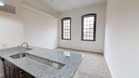 This single-floor loft-style unit showcases a compelling blend of industrial heritage and contemporary upgrades. The open-concept living and kitchen area is anchored by soaring ceilings with exposed ductwork and warm wooden support columns that nod to the building's historic origins. Pendant lighting hangs above a granite-topped peninsula that separates the kitchen from the living space, offering both a functional prep surface and a casual seating area. The kitchen is well-appointed with dark espresso cabinetry extending to the upper reaches of the tall walls, stainless steel appliances including a refrigerator, microwave, and range, and generous counter space throughout. Large, arched multi-pane windows with dark frames flood the living area with natural light and frame views of the surrounding urban streetscape, reinforcing the building's converted warehouse character. Soft, neutral carpet runs through the living and bedroom areas, providing a comfortable contrast to the harder industrial elements. The bedroom offers ample storage with dual bifold closet doors and built-in shelving, set beneath the same impressive ceiling height. The bathroom is clean and straightforward, featuring a full tub-shower combination with white tile surround. Overall, the unit delivers a spacious, light-filled environment with distinctive architectural character throughout.