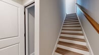 This thoughtfully designed three-story home offers a well-organized layout spread across three distinct levels, each contributing approximately equal floor area to the overall living space. The ground floor includes an attached garage with a motorized door opener and concrete flooring, providing secure, convenient parking and storage. Hardwood-tread staircases with white risers and warm wooden handrails connect each level, giving the home a clean, contemporary feel throughout. The main living level features an open-concept living room with light hardwood flooring and a ceiling fan, flowing seamlessly into a well-appointed kitchen. The kitchen is highlighted by a large center island with a white quartz countertop and built-in sink, complemented by white shaker-style cabinetry, a subway tile backsplash, stainless steel appliances, and a sliding glass door that invites natural light. The upper floor houses the spacious primary bedroom, carpeted for comfort and featuring vaulted ceiling accents, a window, double-door closet, and access to a dedicated walk-in closet with wire shelving. Two bathrooms serve the home, including one with light blue cabinetry, white countertops, and warm hardwood flooring. Neutral tones and clean finishes throughout lend the home a bright, move-in-ready appeal.
