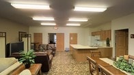 This single-floor space features an open-plan layout that seamlessly integrates a living area and kitchen into one cohesive environment. The living area is furnished with dark brown leather sofas and armchairs arranged around a central coffee table, complemented by patterned carpeting with earthy tones that adds warmth to the space. A decorative green plant provides a natural accent near the seating area, while a small wooden side table adds functional detail.

The kitchen occupies the right side of the space and is equipped with light oak cabinetry, a built-in microwave, a range with stovetop, a refrigerator, and a double sink set into a countertop peninsula. The peninsula extends outward, creating a natural division between the kitchen and living areas while also offering additional workspace. Adjacent dining seating with wooden chairs is positioned near the kitchen for convenient access.

Fluorescent lighting fixtures are evenly distributed across the textured ceiling, providing consistent illumination throughout. Multiple interior doors are visible along the back wall, suggesting access to additional rooms or hallways. An emergency exit sign is prominently displayed, indicating this space is designed with safety compliance in mind.