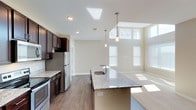 This single-floor residence showcases a thoughtfully designed open-concept layout that seamlessly connects the kitchen and living areas. The kitchen is appointed with rich, dark espresso cabinetry that contrasts elegantly with light granite countertops featuring subtle veining. A central island with an integrated sink and pendant lighting serves as both a functional workspace and a natural gathering point. Stainless steel appliances, including a refrigerator and range, complete the modern culinary space, all set atop warm hardwood-style flooring.

The adjoining living area draws the eye upward with impressively soaring ceilings and a generous bank of windows that bathe the space in abundant natural light. The elevated ceiling height creates a grand sense of openness and airiness rarely found in single-floor living. Neutral, warm-toned walls throughout provide a versatile backdrop well-suited to a variety of interior styles.

Recessed lighting complements the natural illumination, ensuring the space remains bright and welcoming at all hours. The transition from hard flooring to carpet in the living area adds a layer of comfort and definition between spaces. Overall, this home delivers a clean, contemporary aesthetic combined with functional design and a light-filled, airy ambiance.