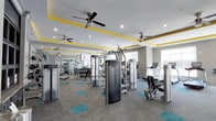 This fitness center is designed to provide a comprehensive workout environment within a single-level layout. The space is outfitted with a robust selection of Life Fitness strength-training machines, a free weight area with a full dumbbell rack, and multiple cardio machines including treadmills, offering options for a wide range of exercise routines. Full-length mirrors line one wall, enhancing the sense of depth and allowing users to monitor their form during workouts. A flat-screen TV is mounted on the wall, adding an entertainment element to training sessions. The ceiling features a distinctive tray design accented with bold yellow trim, adding a vibrant, modern aesthetic to the space. Multiple ceiling fans and recessed lighting ensure the room remains well-lit and comfortably ventilated. The flooring consists of neutral gray carpet tiles punctuated by teal patterned inserts, complementing the room's color scheme. A window along one wall allows natural light to filter in, contributing to an open and energizing atmosphere. Overall, the space is thoughtfully designed to balance functionality with a visually engaging environment.