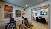 This single-floor business center offers a well-appointed workspace designed to meet a variety of professional needs. The main work area features a built-in white desk with cabinet storage below and an open shelving unit above, providing ample space for books, décor, and personal items. A desktop computer workstation is set up and ready for use, complemented by ergonomic task chairs and additional patterned seating for a comfortable yet functional atmosphere. The walls are adorned with eclectic, typographic artwork that adds a creative and contemporary character to the space. Neutral, light-toned walls and recessed lighting create a bright, focused environment conducive to work. An open doorway leads directly into an adjoining conference room, which is furnished with a large meeting table surrounded by rolling office chairs — ideal for collaborative sessions or small group meetings. The conference room continues the same clean, modern aesthetic with additional artwork on the walls. The cohesive design throughout, combining practical furnishings with thoughtful decorative elements, makes this business center both a functional and visually engaging professional environment.