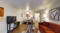 This single-floor residence offers a thoughtfully designed open-concept layout that seamlessly connects the living, dining, and kitchen areas. The living room is anchored by rich hardwood-style flooring that extends throughout the main living space, creating a warm and cohesive aesthetic. A ceiling fan provides both comfort and a subtle design element overhead, while clean white walls offer a neutral backdrop that enhances the sense of openness and light throughout.

The living area comfortably accommodates a sofa and media console, making it well-suited for relaxing or entertaining. Adjacent to the living space, the dining area features a charming dining set positioned beneath a second ceiling fan, ideal for everyday meals or casual gatherings. Colorful accent pieces and decorative artwork add personality and vibrancy to the space.

A raised kitchen bar separates the dining area from the kitchen, maintaining an open flow while defining each functional zone. The kitchen is partially visible beyond the bar, offering a practical pass-through design. A hallway leading from the main living area suggests additional rooms beyond, completing the layout of this well-appointed single-floor home. Overall, the space strikes a balance between functional design and inviting, comfortable living.