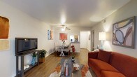 This single-floor residence offers a thoughtfully designed open-concept layout that seamlessly connects the living, dining, and kitchen areas. The living room is anchored by rich hardwood-style flooring that extends throughout the main living space, creating a warm and cohesive aesthetic. A ceiling fan provides both comfort and a subtle design element overhead, while clean white walls offer a neutral backdrop that enhances the sense of openness and light throughout.

The living area comfortably accommodates a sofa and media console, making it well-suited for relaxing or entertaining. Adjacent to the living space, the dining area features a charming dining set positioned beneath a second ceiling fan, ideal for everyday meals or casual gatherings. Colorful accent pieces and decorative artwork add personality and vibrancy to the space.

A raised kitchen bar separates the dining area from the kitchen, maintaining an open flow while defining each functional zone. The kitchen is partially visible beyond the bar, offering a practical pass-through design. A hallway leading from the main living area suggests additional rooms beyond, completing the layout of this well-appointed single-floor home. Overall, the space strikes a balance between functional design and inviting, comfortable living.