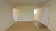 This single-floor residence offers a clean, functional layout with open, flowing living areas designed to maximize usability. The main living space is finished with wall-to-wall carpet in a warm neutral tone, complemented by crisp white walls and white baseboards that give the interior a bright, airy feel. A central dividing wall with open passageways connects the primary living area to an adjacent room, creating a sense of continuity while still defining separate spaces. Natural light filters in through a window in the adjoining area, further enhancing the open atmosphere. A floor-level air vent is built into the base of the central wall, indicating a dedicated HVAC system for climate control throughout. Electrical outlets are conveniently positioned along the walls, and a wall-mounted panel is visible, suggesting integrated utility infrastructure. A small section of hardwood or laminate flooring near the entry provides a practical transition surface. The overall ambiance is understated and versatile, offering a neutral canvas well-suited for a variety of interior design preferences. The straightforward, single-level layout makes the space highly accessible and easy to navigate.
