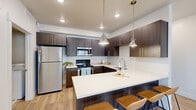 This thoughtfully laid-out single-floor residence offers 1 bedroom and 1 bathroom across a compact yet functional footprint. Light-toned hardwood flooring runs throughout, creating a cohesive and warm aesthetic that unifies every room.

The kitchen is a standout feature, showcasing dark wood-grain cabinetry that extends floor to ceiling, contrasted by crisp white quartz countertops. A large peninsula with seating provides both additional prep space and a casual dining area. Stainless steel appliances—including a refrigerator, electric range, and over-the-range microwave—complete the modern, well-equipped kitchen. Pendant lighting over the peninsula adds a polished finishing touch.

The living room is open and airy, bathed in natural light from a large window fitted with horizontal blinds. Neutral white walls and recessed lighting contribute to a clean, versatile space.

The bedroom is a comfortable, well-proportioned retreat featuring its own window for natural light and a double-door closet offering ample storage.

The bathroom features white quartz countertops, a large vanity mirror, and a clean, minimalist design. A combined bathroom and laundry area includes an in-unit washer and dryer alongside a spacious double-door storage closet, adding significant everyday convenience to this efficiently designed home.