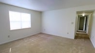 This single-floor space presents a straightforward and functional layout finished with consistent neutral tones throughout. The interior features wall-to-wall carpeting in a warm beige hue, providing a soft and comfortable underfoot feel that extends across the living areas. The walls are painted in a clean, light neutral tone, creating a bright and open atmosphere that pairs well with the textured popcorn ceiling overhead. A window fitted with a white blind allows natural light to filter into the room, further enhancing the airy feel of the space. Electrical outlets are thoughtfully placed along the walls, offering practical convenience throughout. The clean lines and understated finishes give the space a versatile, move-in-ready quality suitable for a variety of furnishing styles. The overall ambiance is calm and uncluttered, making it easy to envision customizing the interior to suit personal preferences. The single-floor design ensures easy accessibility and a cohesive flow throughout the entire space.