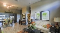 This single-floor residence offers a thoughtfully designed open-concept layout that maximizes both space and functionality. The living area is an immediate focal point, highlighted by dramatically high ceilings and elegant pendant lighting that draws the eye upward. A tufted cream sofa anchors the sitting area, complemented by warm earth-toned accent pillows and a sleek dark coffee table. Botanical-themed framed artwork adorns the walls, adding a fresh, nature-inspired aesthetic throughout. Natural light filters into the space, enhancing the warm, neutral color palette of soft greiges and sage tones that span the walls. Light hardwood-style flooring runs continuously through the main living and dining areas, creating a cohesive and airy feel. The dining area sits just adjacent to the living space, furnished with a dark wood table and chairs, making it ideal for everyday dining or entertaining. A glimpse of stainless steel appliances is visible beyond, hinting at a well-appointed kitchen nearby. A hallway leads further into the residence, suggesting additional private rooms beyond. Overall, the space strikes a balance between modern sophistication and comfortable, livable warmth.