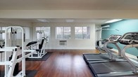 This fitness center is a thoughtfully designed single-level space built to support a well-rounded exercise routine. The room features warm hardwood flooring throughout, providing a clean and durable surface suited for a variety of workout activities. Flush-mounted ceiling light panels ensure consistent, even illumination across the space, while large windows fitted with vertical blinds along one wall admit natural light and offer views of the surrounding outdoor greenery.

A bold teal accent wall anchors one end of the room, adding visual depth and contrast to the otherwise neutral cream-colored walls. Full-length mirrors are strategically positioned to reflect both light and the layout of equipment, enhancing the sense of openness throughout the space.

The fitness center is outfitted with multiple Paramount strength training machines, providing options for a range of resistance exercises. Cardio equipment includes several treadmills and an elliptical trainer. Protective rubber mats are placed beneath each piece of equipment to safeguard the flooring and provide stability during use. A wall-mounted air conditioning unit supports climate control, ensuring a comfortable environment year-round. The entry door opens directly to an outdoor walkway, allowing for easy ventilation and access.