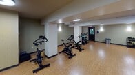 Located at 3121 S Chowen Ave in Minneapolis, Minnesota, this single-floor fitness studio offers a well-equipped environment designed for group exercise and individual training. The space features warm wood-style flooring throughout, providing a durable yet inviting surface suited for a variety of workout activities.

The cycling area is a central highlight, housing multiple stationary spin bikes arranged for group classes. Structural columns are integrated into the layout, defining zones within the open floor plan without significantly obstructing usable space. Wall-mounted sconces provide ambient lighting alongside flush ceiling fixtures, creating a consistently lit environment throughout.

Full-length mirrors line portions of the walls, enhancing the sense of openness and allowing users to monitor their form during workouts. A large flat-screen display is mounted prominently in one section of the room, visible from across the floor and suitable for streaming fitness content or class programming.

Additional accessories such as stability balls are present, indicating the space supports varied fitness modalities beyond cycling. Private rooms with doors are accessible from the main area, likely serving as changing or storage spaces. Emergency exit signage is clearly posted, reflecting adherence to safety standards. Overall, the studio presents a functional and organized fitness environment.