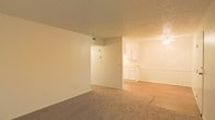This single-floor space features an open-concept layout that connects the main living area to the kitchen and dining zone. The living area is finished with wall-to-wall carpet in a neutral tone, providing a comfortable and cohesive foundation throughout the space. A textured ceiling runs across the entirety of the unit, adding subtle dimension overhead.

A partial dividing wall separates the living area from the kitchen and dining space, maintaining a sense of openness while defining each functional zone. The transition from carpet to hard-surface flooring marks the shift into the kitchen area, where white cabinetry lines the walls, offering ample storage. A ceiling-mounted light fixture illuminates the dining and kitchen zone, complementing the warm, ambient tones of the space.

The walls throughout are painted in a clean, neutral tone, lending a blank-canvas quality ideal for personalization. A visible HVAC vent is integrated into the upper wall, ensuring climate control throughout. Overall, the space presents a practical and straightforward layout with consistent finishes and a warm, understated aesthetic.