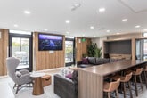 Located at 185 N West Temple in Salt Lake City, Utah, this single-floor space offers a thoughtfully designed open layout that seamlessly blends a living room and kitchenette into one cohesive environment. The interior is defined by a clean, contemporary aesthetic, featuring light-toned large-format tile flooring, crisp white ceilings with recessed lighting, and warm wood-grain accent panels that add texture and visual depth throughout.

The spacious living area is anchored by a generously sized sectional sofa and a plaid ottoman, creating an inviting gathering space. Built-in wall niches with accent lighting flank the room, providing both architectural interest and functional display areas. A wall-mounted television adds a media-ready element to the lounge setting. Tall indoor plants introduce a refreshing natural element, complementing the neutral color palette.

The kitchenette is efficiently laid out along a single wall, featuring warm wood cabinetry, a white quartz countertop, an undermount sink, a compact oven, and beverage refrigerators. Open floating shelves with under-cabinet lighting display décor and everyday essentials with style.

A striking wood-clad structural column anchors the center of the space, reinforcing the modern design language. Glass doors open directly to an outdoor patio, allowing natural light to fill the interior and offering a seamless indoor-outdoor connection with views of the surrounding urban environment.