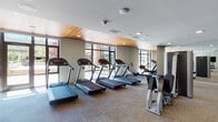 Located at 140 SW Columbia St in Portland, Oregon, this well-appointed fitness center offers a comprehensive workout environment designed to accommodate a wide range of exercise needs. The facility is organized across a single floor with distinct zones for cardio, strength training, and functional fitness.

The main cardio area is a standout feature, lined with floor-to-ceiling windows that flood the space with natural light and provide views of the surrounding Portland streetscape. Multiple treadmills, elliptical machines, and stationary bikes are arranged along the window wall, creating an open and energizing workout environment. A warm wood-paneled ceiling accent adds a refined architectural touch to this section.

Adjacent to the cardio area, a dedicated strength training zone houses a full complement of cable machines, selectorized equipment, and multi-functional machines. Free weight stations are well-stocked with dumbbells and kettlebells in graduated sizes, accompanied by adjustable benches for a variety of exercises.

A separate functional training room provides space for dynamic workouts, equipped with medicine balls, stability balls, resistance tools, and foldable mats stored on organized racks. A large wall-mounted display screen and curated wall art contribute to the room's motivating atmosphere.

Throughout the facility, neutral-toned carpet tile flooring, recessed lighting, and a clean, modern aesthetic create a professional and welcoming environment suited for focused training.