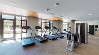 Located at 140 SW Columbia St in Portland, Oregon, this well-appointed fitness center offers a comprehensive workout environment designed to accommodate a wide range of exercise needs. The facility is organized across a single floor with distinct zones for cardio, strength training, and functional fitness.

The main cardio area is a standout feature, lined with floor-to-ceiling windows that flood the space with natural light and provide views of the surrounding Portland streetscape. Multiple treadmills, elliptical machines, and stationary bikes are arranged along the window wall, creating an open and energizing workout environment. A warm wood-paneled ceiling accent adds a refined architectural touch to this section.

Adjacent to the cardio area, a dedicated strength training zone houses a full complement of cable machines, selectorized equipment, and multi-functional machines. Free weight stations are well-stocked with dumbbells and kettlebells in graduated sizes, accompanied by adjustable benches for a variety of exercises.

A separate functional training room provides space for dynamic workouts, equipped with medicine balls, stability balls, resistance tools, and foldable mats stored on organized racks. A large wall-mounted display screen and curated wall art contribute to the room's motivating atmosphere.

Throughout the facility, neutral-toned carpet tile flooring, recessed lighting, and a clean, modern aesthetic create a professional and welcoming environment suited for focused training.